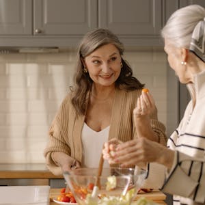 Two senior women smiling and preparing a fresh salad in a cozy kitchen setting, showcasing friendship and healthy living.